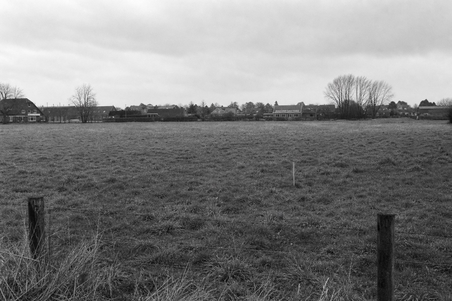 Leeg grasveld met een houten meetpaal achter een draadafrastering aan de rand van een Nederlands dorp